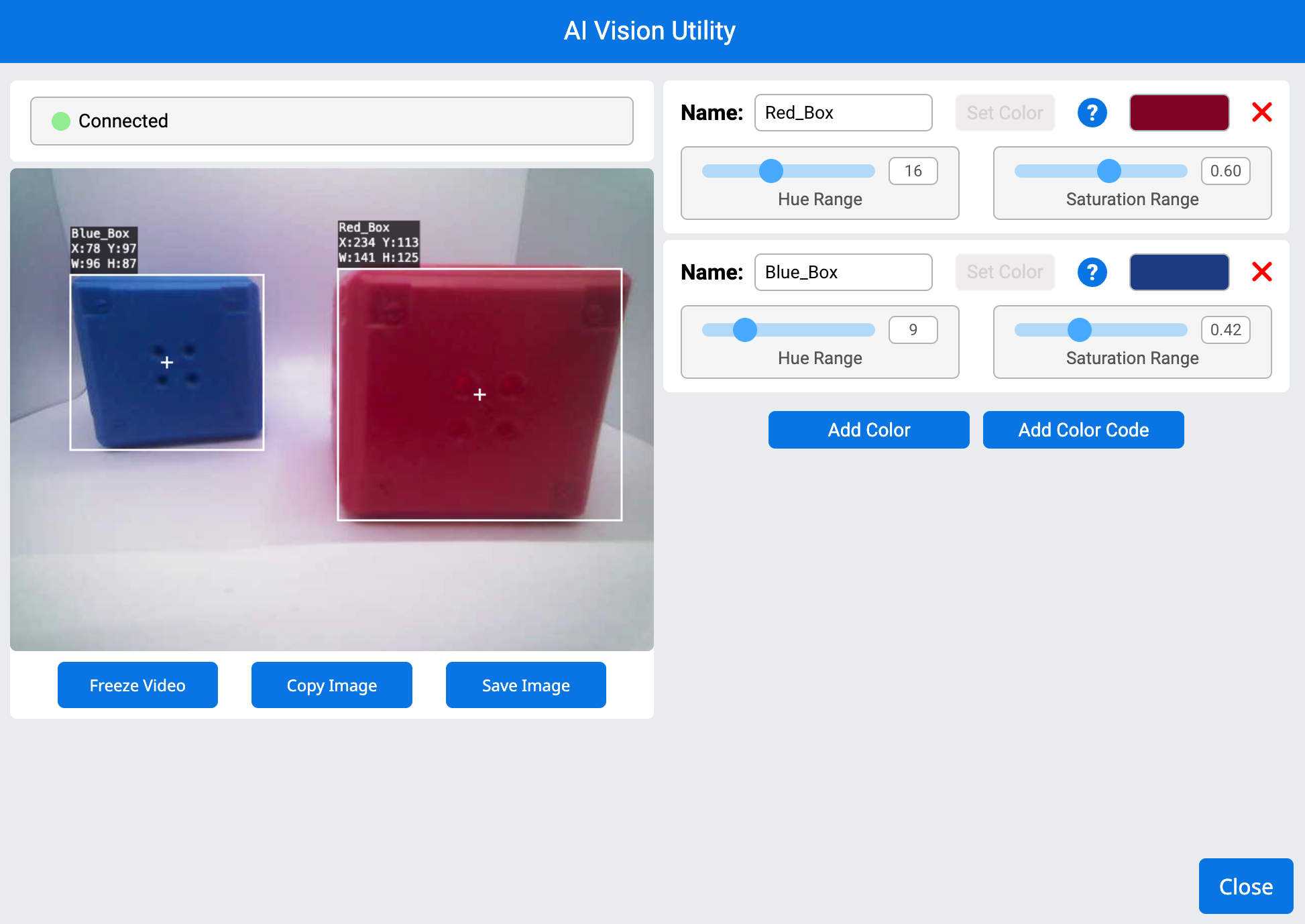 The AI Vision Utility showing a connected vision sensor detecting two colored objects. The left side displays a live camera feed with a blue box on the left and a red box on the right, each outlined with white bounding boxes. Black labels display their respective names, coordinates, and dimensions. The right side contains color signature settings, with sliders for hue and saturation range for both the red and blue boxes. Buttons for adding colors, freezing video, copying, and saving the image are at the bottom, along with a close button in the lower right corner.